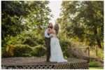 Groom dips and kisses his bride in a romantic moment on a wooden platform surrounded by lush greenery, with the bride holding a bouquet of pink and white flowers.