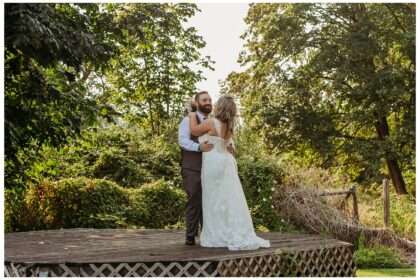 Groom dips and kisses his bride in a romantic moment on a wooden platform surrounded by lush greenery, with the bride holding a bouquet of pink and white flowers.