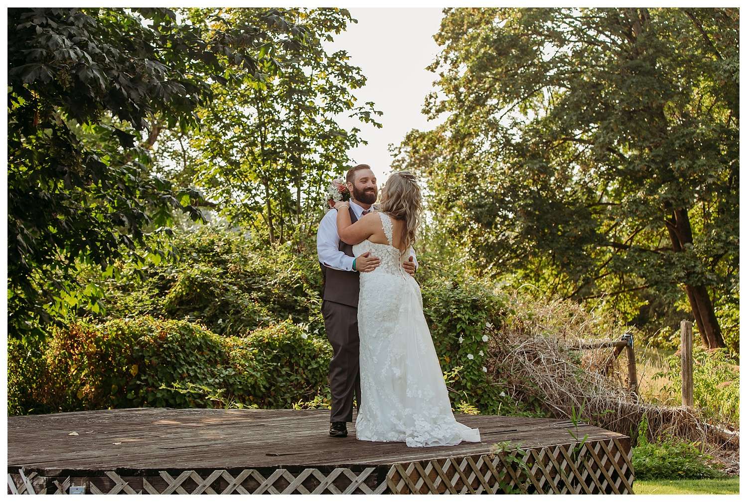 Groom dips and kisses his bride in a romantic moment on a wooden platform surrounded by lush greenery, with the bride holding a bouquet of pink and white flowers.