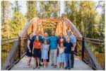 Extended family of eleven poses on a wooden bridge at Bothell Landing in Bothell, Washington, surrounded by tall green trees. The group includes grandparents, parents, and five children, all smiling and dressed in coordinated, colorful outfits on a sunny day.