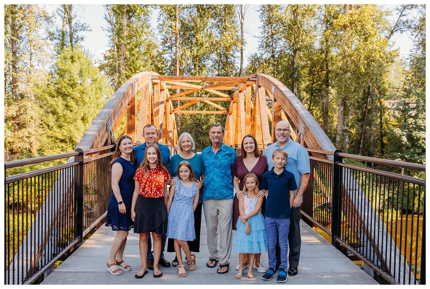 Extended family of eleven poses on a wooden bridge at Bothell Landing in Bothell, Washington, surrounded by tall green trees. The group includes grandparents, parents, and five children, all smiling and dressed in coordinated, colorful outfits on a sunny day.