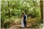 Bride and groom share a kiss in a sun-dappled forest at Nature’s Connection in Arlington, WA. The bride holds a colorful bouquet and wears a lace dress, while the groom is dressed in a navy vest and slacks, surrounded by lush greenery and tall trees.