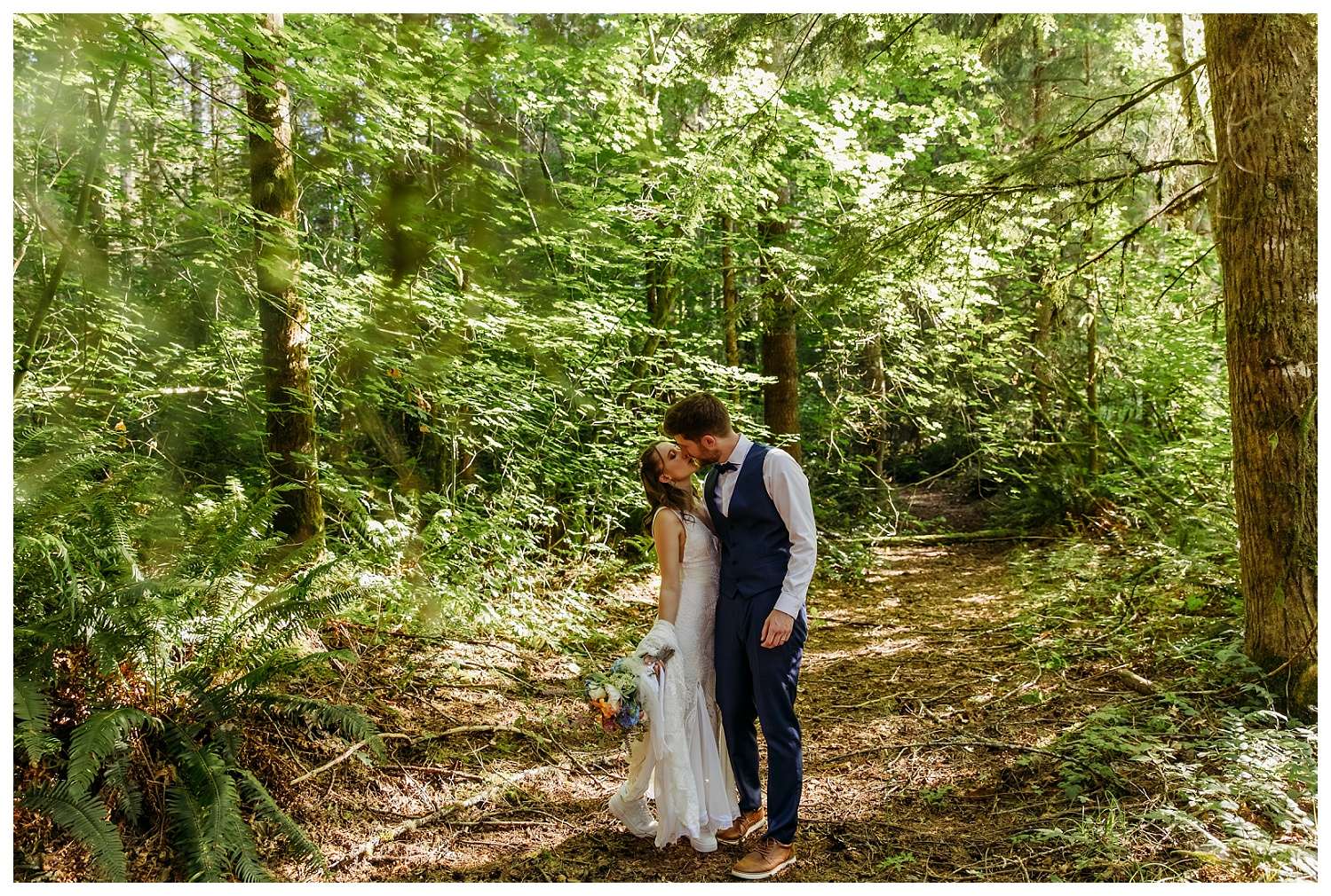Bride and groom share a kiss in a sun-dappled forest at Nature’s Connection in Arlington, WA. The bride holds a colorful bouquet and wears a lace dress, while the groom is dressed in a navy vest and slacks, surrounded by lush greenery and tall trees.