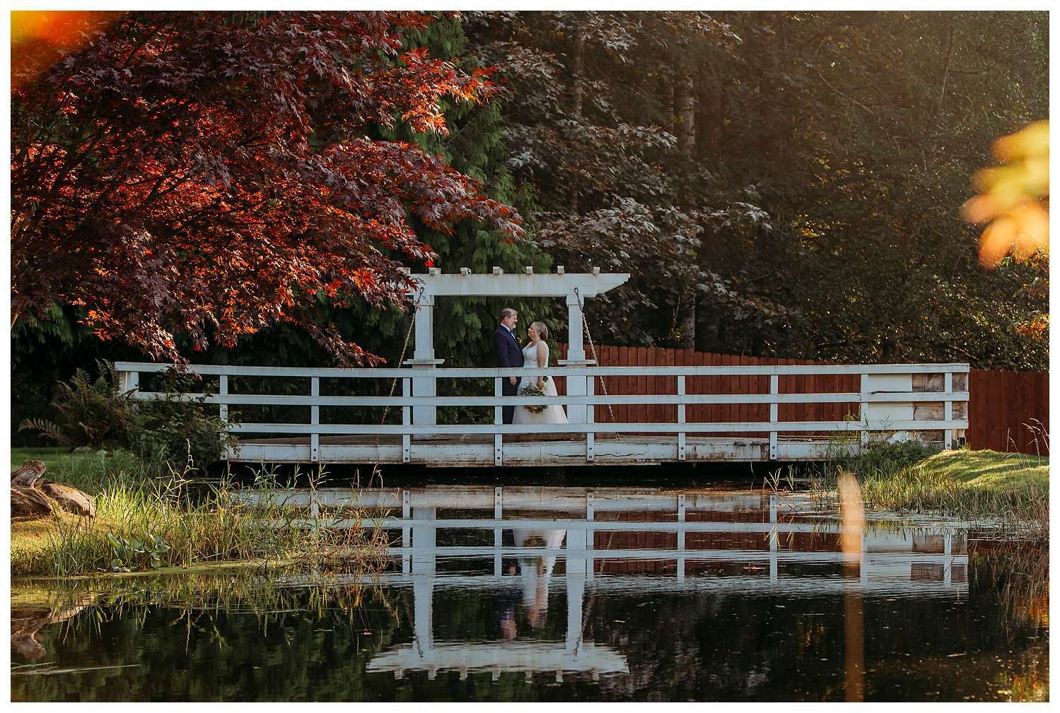 Bride and groom stand on a white wooden bridge over a reflective pond, surrounded by vibrant summer foliage and golden light at Nature’s Connection in Arlington, WA.