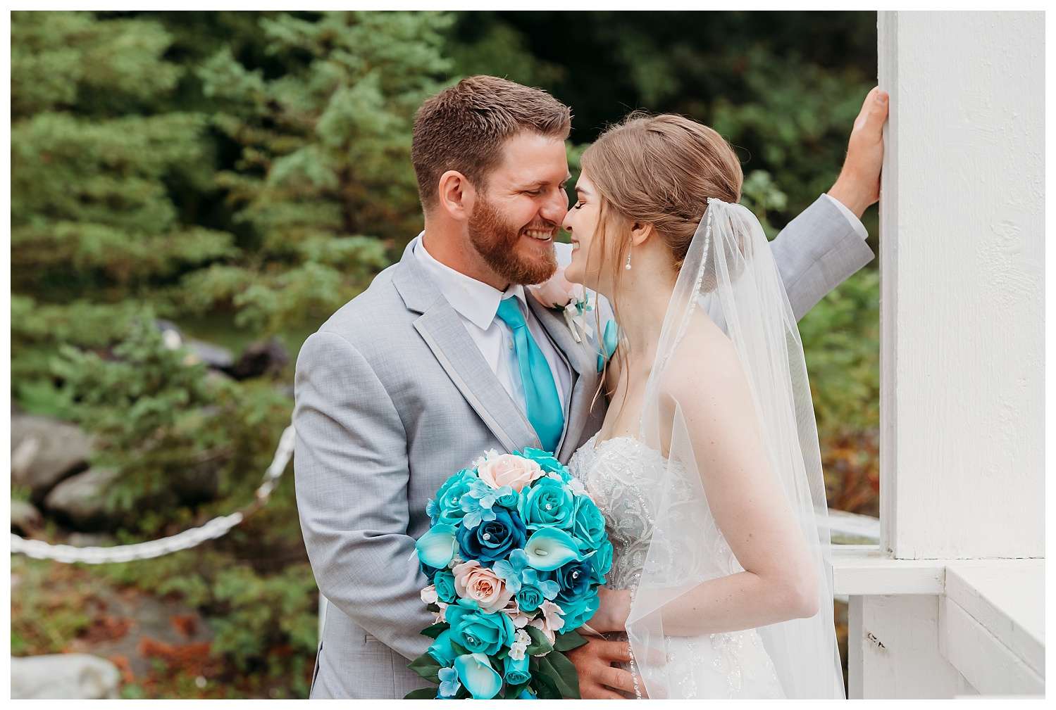 Bride and groom sharing an intimate moment on a white wooden bridge surrounded by greenery, with the bride holding a cascading bouquet of turquoise, blush, and white flowers.