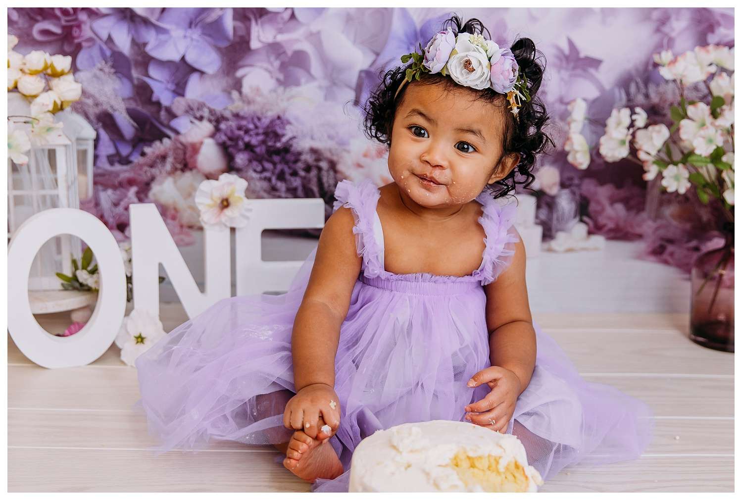 One-year-old girl celebrating her birthday in a whimsical purple-themed cake smash, surrounded by balloons and butterfly decorations, wearing a lavender tutu and butterfly headband, joyfully smashing a purple ombré cake with frosting-covered hands and a big smile.