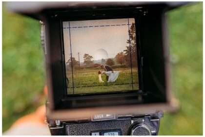 A photo taken through the viewfinder of a vintage film camera shows a groom dipping his bride for a kiss in a flower-filled field at sunset. The romantic scene is framed within the camera, highlighting the rustic charm of Terra Valley Farms. Title: Blooms, Booms, and a Love in Full Harvest: A September Wedding at Terra Valley Farms.