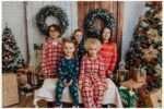 A smiling family of four poses in matching holiday pajamas inside a festive studio decorated for Christmas, featuring wreaths, Christmas trees with ornaments and lights, wrapped presents, and a "Letters to Santa" box.