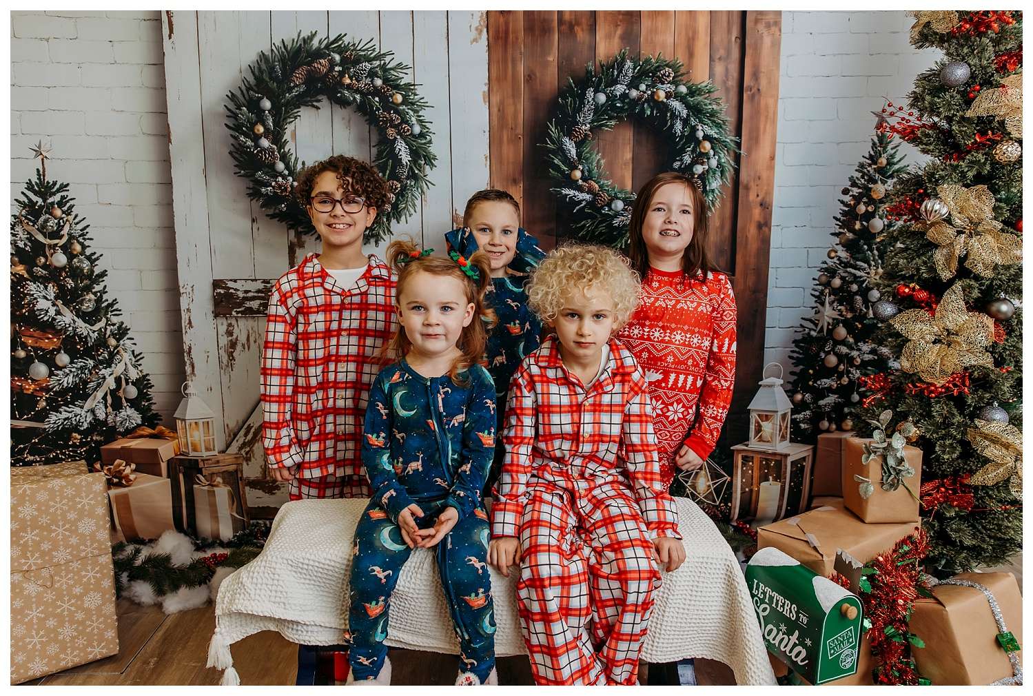 A smiling family of four poses in matching holiday pajamas inside a festive studio decorated for Christmas, featuring wreaths, Christmas trees with ornaments and lights, wrapped presents, and a "Letters to Santa" box.