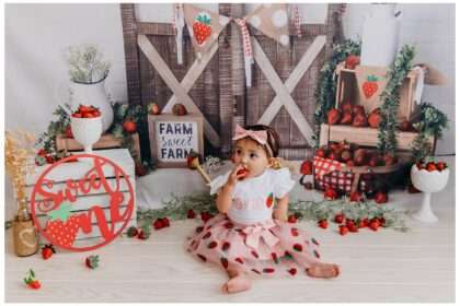 One-year-old girl celebrating her strawberry-themed cake smash with pink frosting, strawberry decor, and bright red and green accents in a photo studio.