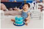 One-year-old baby boy in a blue romper sitting in front of a nautical-themed backdrop with sailboats, seashells, and the word “ONE” in white letters.