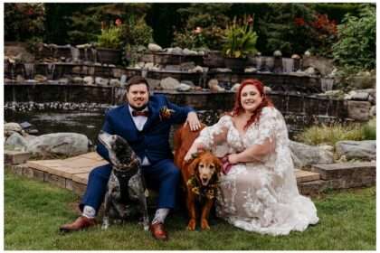Bride and groom seated outdoors by a cascading stone waterfall, smiling with their two dogs—one golden retriever wearing a floral collar and one black and white dog with a bowtie harness—on their wedding day at Nature’s Connection.