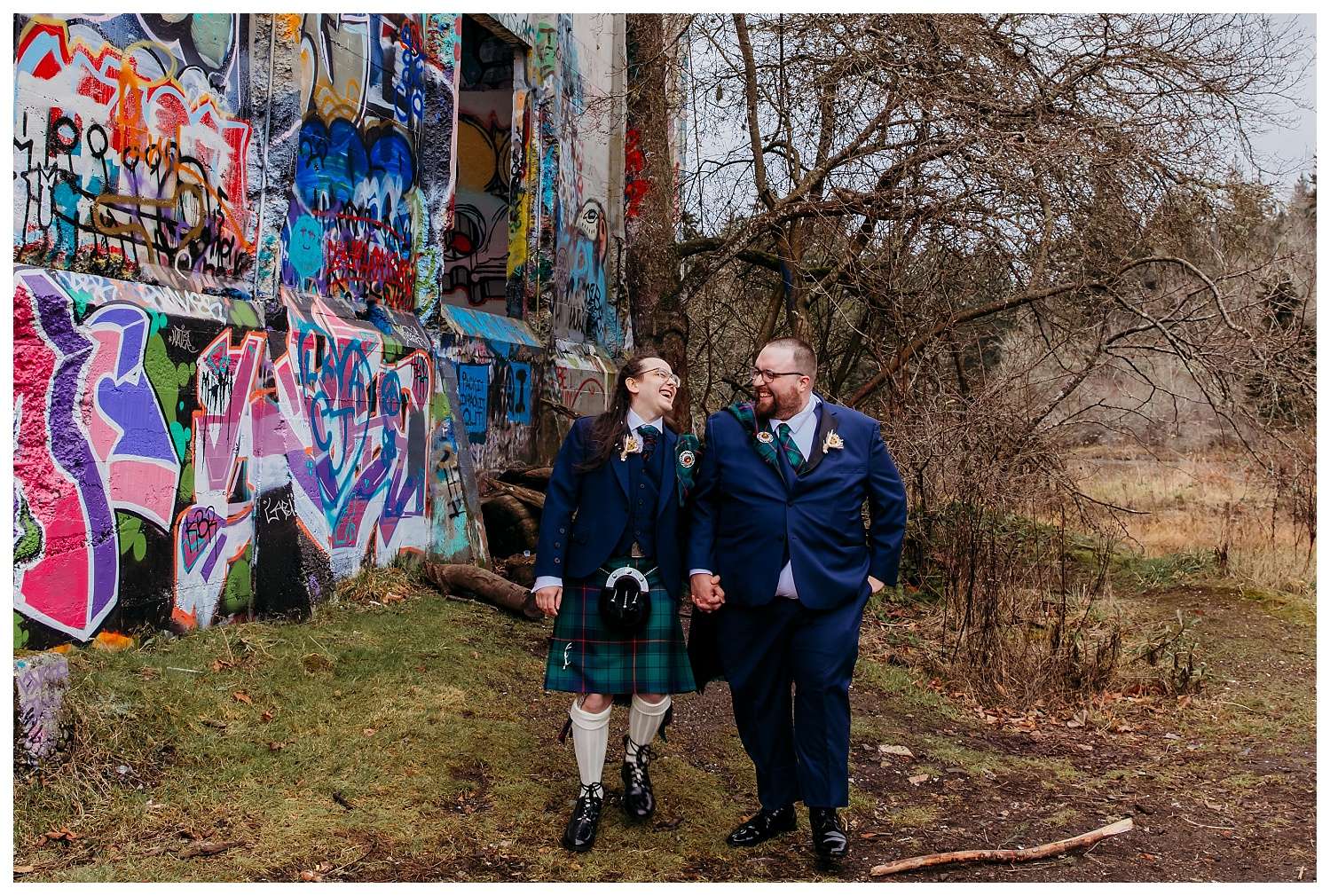 Two grooms wearing traditional Scottish kilts, sashes, and sporrans stand together joyfully after their wedding ceremony at Bainbridge Island Municipal Court, with post-ceremony portraits taken at the scenic Blakely Harbor Park surrounded by trees and waterfront views.