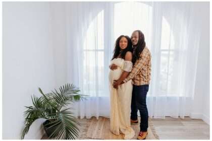 Pregnant woman in a cream lace maternity gown stands beside her partner in a bright, airy studio with large arched windows and sheer white curtains; her partner gently embraces her from behind as they both smile softly.