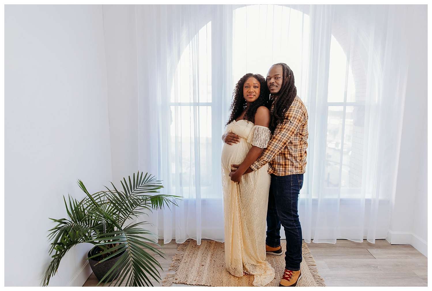 Pregnant woman in a cream lace maternity gown stands beside her partner in a bright, airy studio with large arched windows and sheer white curtains; her partner gently embraces her from behind as they both smile softly.