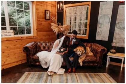 Bride and groom sit on a vintage leather couch with their golden retriever at Pine Creek Nursery. The bride leans affectionately toward the groom while holding a bouquet of roses and greenery. The groom smiles as he pets their dog, who wears a navy bow tie. The cozy indoor setting features wood-paneled walls, pampas grass decor, and a macramé wall hanging, capturing a warm, rustic vibe perfect for a rainy September wedding day.