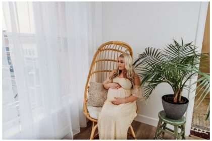 Pregnant woman in a cream lace gown sits peacefully in a rattan chair by a window with sheer curtains, gently cradling her belly beside a tall potted palm plant in a cozy, sunlit studio.