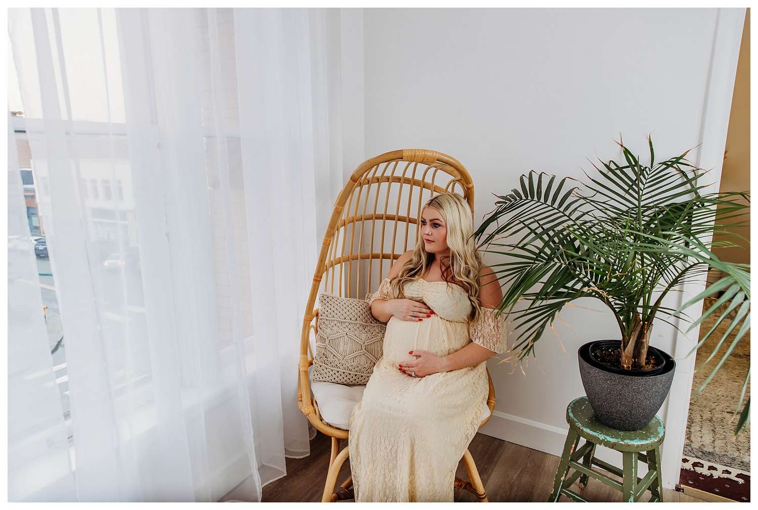 Pregnant woman in a cream lace gown sits peacefully in a rattan chair by a window with sheer curtains, gently cradling her belly beside a tall potted palm plant in a cozy, sunlit studio.