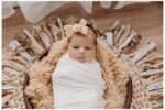 Three-week-old baby girl Madison swaddled in a creamy white wrap, lying in a cozy brown bowl lined with a fluffy tan blanket, wearing a beige lace bow headband and surrounded by rustic, textured decor on a wooden floor.