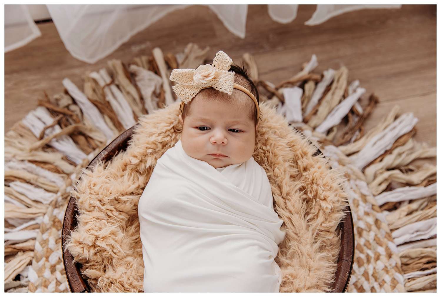 Three-week-old baby girl Madison swaddled in a creamy white wrap, lying in a cozy brown bowl lined with a fluffy tan blanket, wearing a beige lace bow headband and surrounded by rustic, textured decor on a wooden floor.