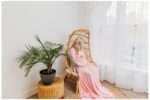 "Pregnant woman in a fitted pink gown with a flowing train, sitting gracefully in a woven boho egg chair against white studio walls, softly lit in a bright, airy downtown Everett photo studio."