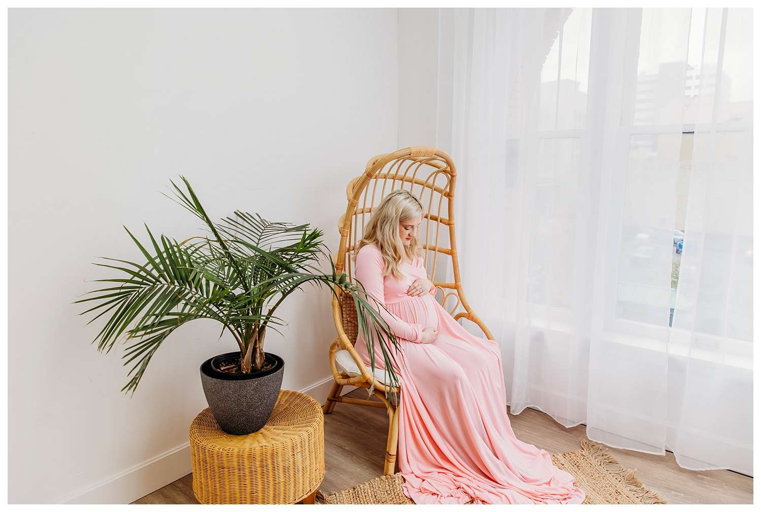"Pregnant woman in a fitted pink gown with a flowing train, sitting gracefully in a woven boho egg chair against white studio walls, softly lit in a bright, airy downtown Everett photo studio."
