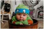 Baby dressed as a Ninja Turtle in a green costume with a blue eye mask, resting inside a rustic wooden barrel. The Halloween-themed setup includes black pumpkins, spider webs, skeleton decorations, and spooky backdrop elements like tombstones and ghostly figures. The baby looks directly at the camera with a curious expression.