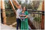 Smiling family of three stands on a wooden bridge at Bothell Landing, holding their 6-month-old baby boy who is dressed in a blue shirt and pants. The mother wears a green floral dress, and the father wears a light gray shirt and navy pants. Lush trees and a calm river are visible in the background.