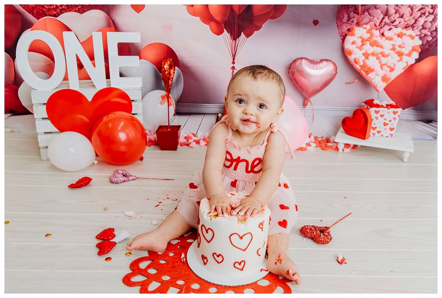 One-year-old girl celebrating her first birthday in a Valentine’s-themed cake smash, surrounded by a heart-shaped air balloon backdrop, wearing a pink outfit with a red bow, joyfully playing with a heart-decorated smash cake in a downtown Everett photo studio.