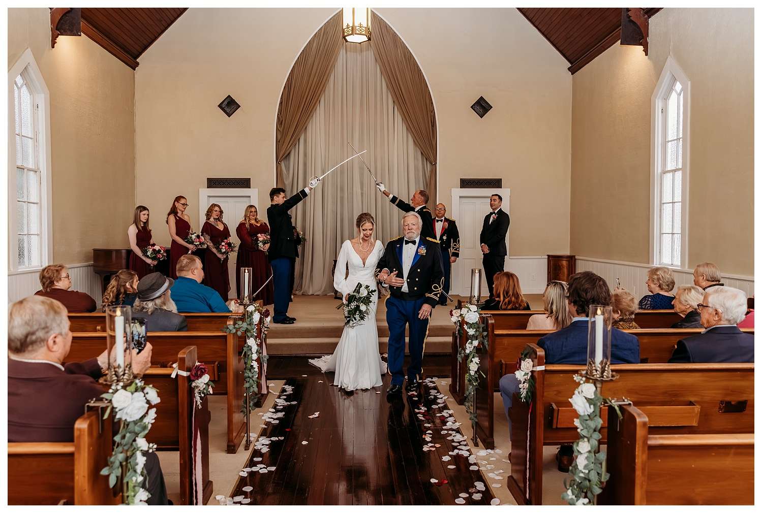 A bride in a long-sleeved white gown walks arm-in-arm with her groom in a formal military dress uniform under a traditional saber arch at Belle Chapel. Bridesmaids in deep burgundy dresses and groomsmen in military attire stand on either side, while guests seated in pews decorated with white and burgundy floral arrangements watch the joyful exit.