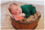 Newborn baby Matthew, 10 days old, peacefully sleeping in a round wooden bucket lined with soft brown wool, wearing a dark green outfit and resting his head on a peach-colored velvet pillow in a cozy studio setting.