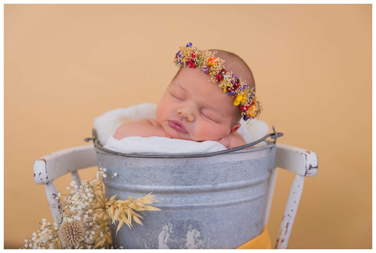Newborn baby peacefully sleeping with a delicate floral crown on her head, swaddled in a soft blanket, surrounded by gentle pastel tones.