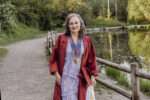 Conchita walks toward the camera on a park path beside a pond, smiling in her graduation gown and medals, surrounded by greenery in soft evening light.