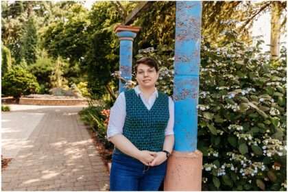 Person wearing glasses and a patterned teal vest over a white shirt stands outdoors at a garden, leaning on a wooden railing with one hand in their pocket, surrounded by trees and greenery.