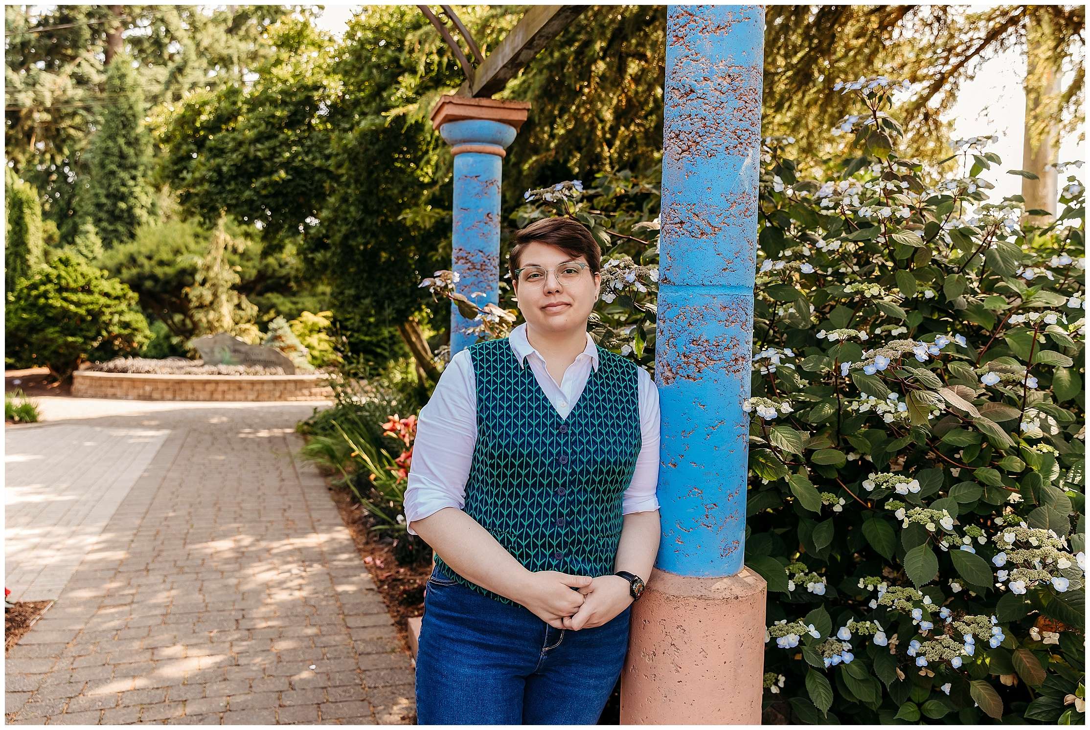 Person wearing glasses and a patterned teal vest over a white shirt stands outdoors at a garden, leaning on a wooden railing with one hand in their pocket, surrounded by trees and greenery.
