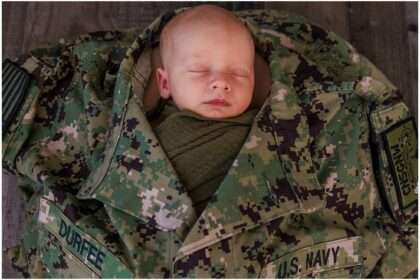 Newborn baby boy wrapped in a green swaddle, sleeping inside a retired U.S. Navy uniform with a woodland theme backdrop.