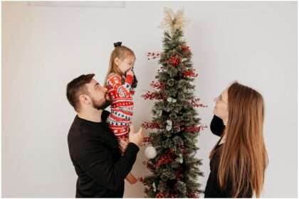 Family of three in matching holiday pajamas decorating a Christmas tree in a downtown Everett photography studio, sharing pregnancy announcement ultrasound photo during Christmas session.