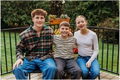 Family of five smiling together outdoors in their front yard during autumn with golden November light and evergreen trees in the background.