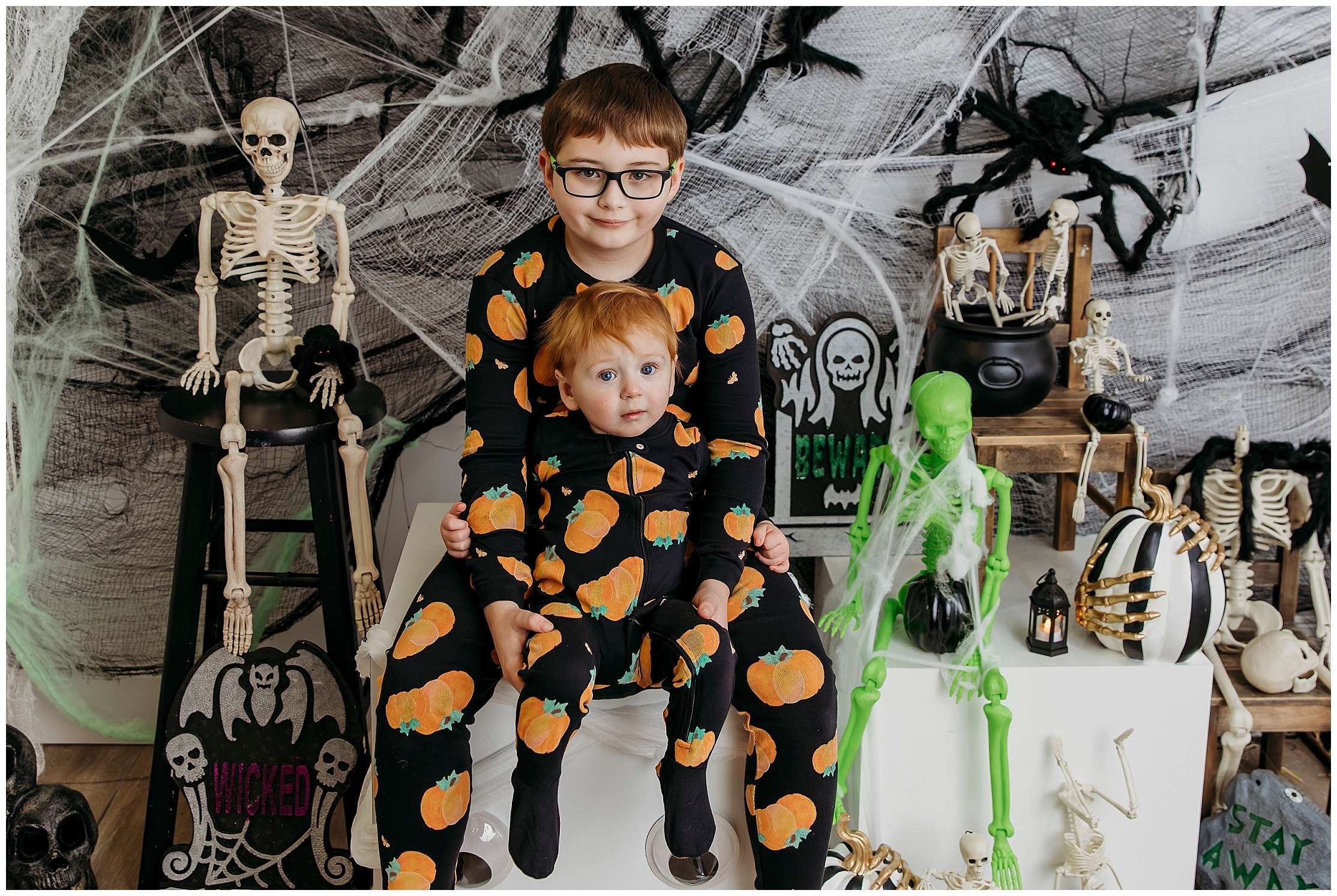 Two young children in matching pumpkin pajamas sit together surrounded by Halloween skeleton decorations, cobwebs, and spooky props during a themed studio photo session.