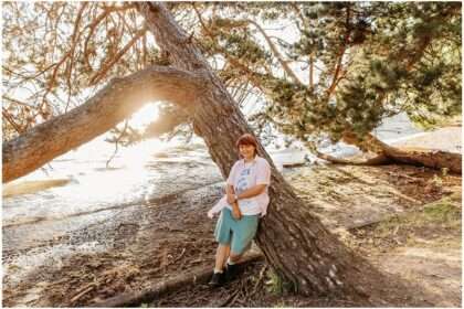 High school senior leaning against a curved tree at Denny Park during golden hour with sunlight shining through branches by the water