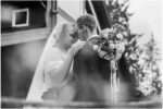Black and white photo of bride and groom embracing closely during an outdoor wedding, with the bride holding a bouquet featuring a sentimental locket.