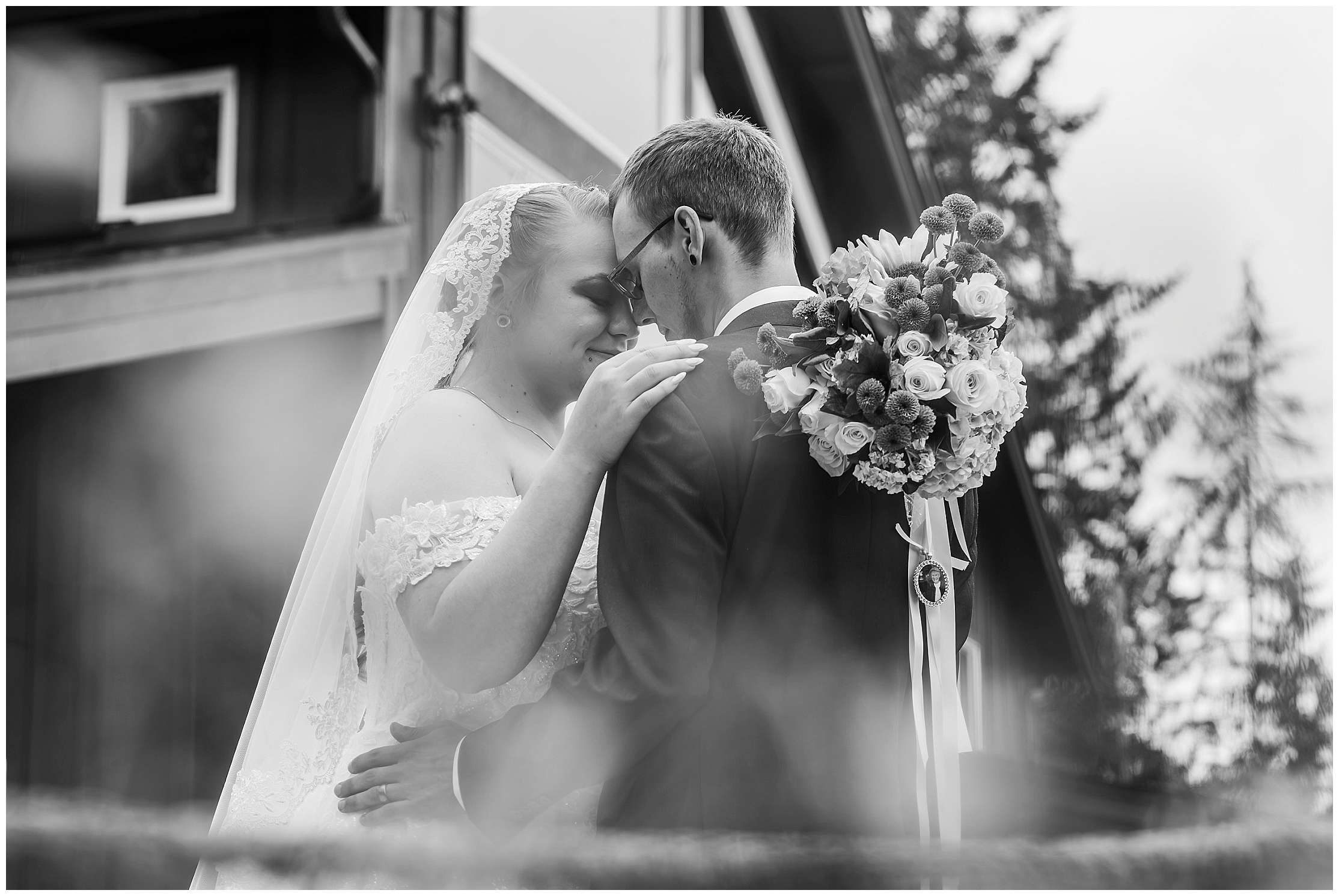 Black and white photo of bride and groom embracing closely during an outdoor wedding, with the bride holding a bouquet featuring a sentimental locket.