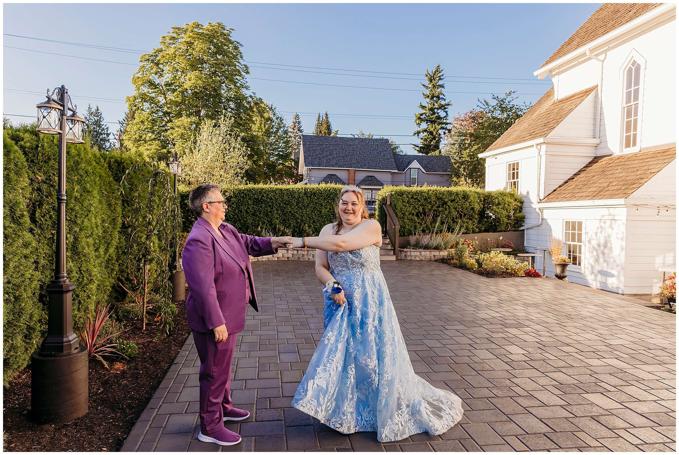 Two brides share a joyful moment outside Snohomish Belle Chapel, one in a lavender suit twirling her partner in a light blue lace wedding gown on a sunlit brick courtyard surrounded by greenery.