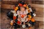 Newborn baby in pumpkin outfit surrounded by fall flowers and pumpkins in a boho Halloween-themed studio setup