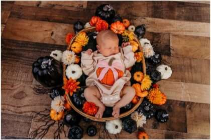 Newborn baby in pumpkin outfit surrounded by fall flowers and pumpkins in a boho Halloween-themed studio setup