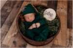 Newborn baby Silas sleeping in a wooden bowl wearing a green outfit and bonnet, surrounded by soft green textures in a natural-themed studio setup
