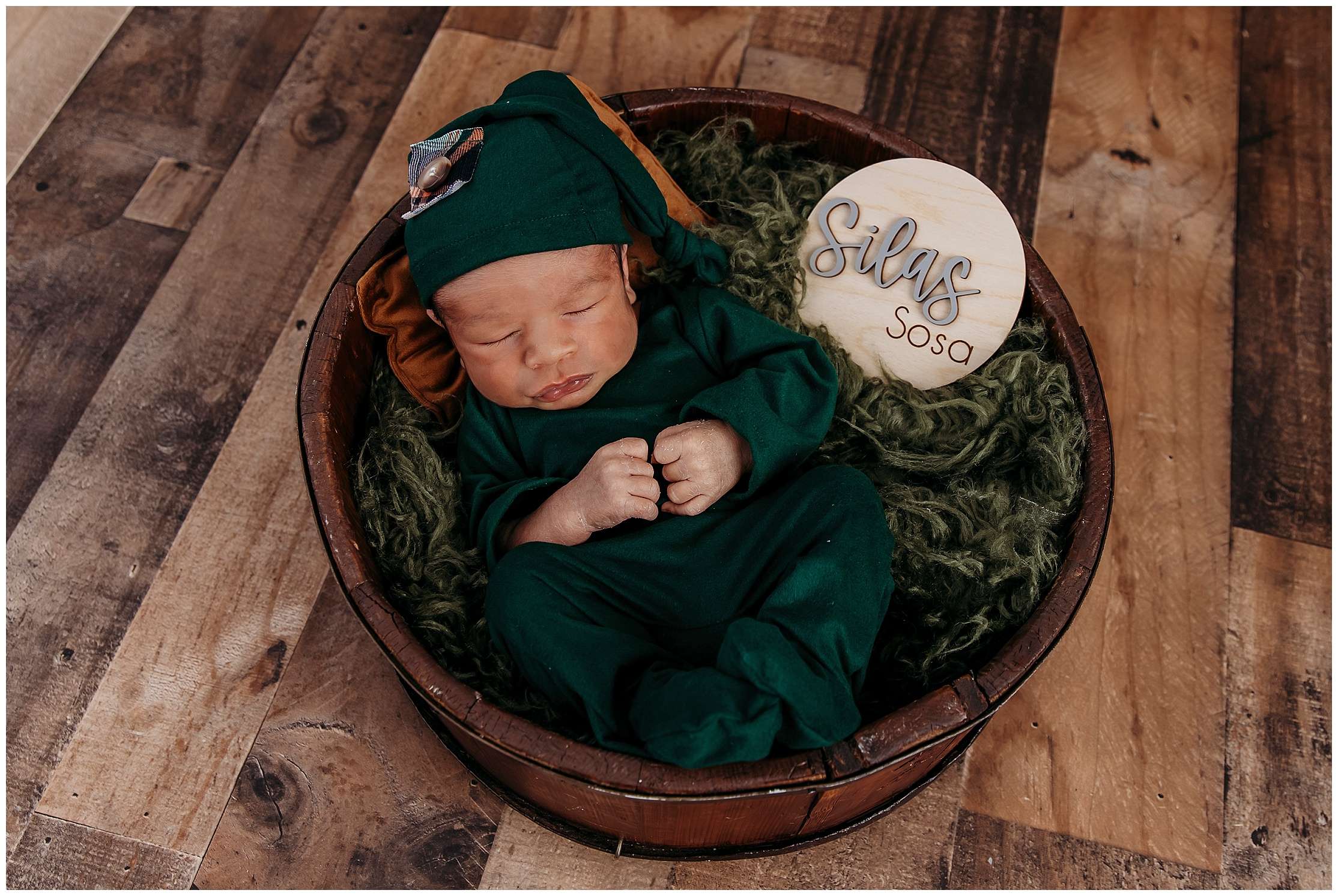 Newborn baby Silas sleeping in a wooden bowl wearing a green outfit and bonnet, surrounded by soft green textures in a natural-themed studio setup