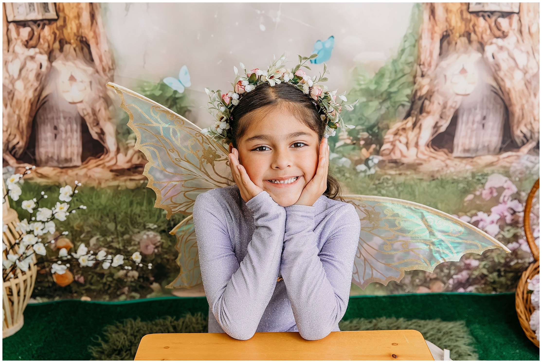 A smiling 7-year-old girl wearing gold fairy wings and a flower crown holds a wand in a woodland-themed studio setup filled with soft, magical details.