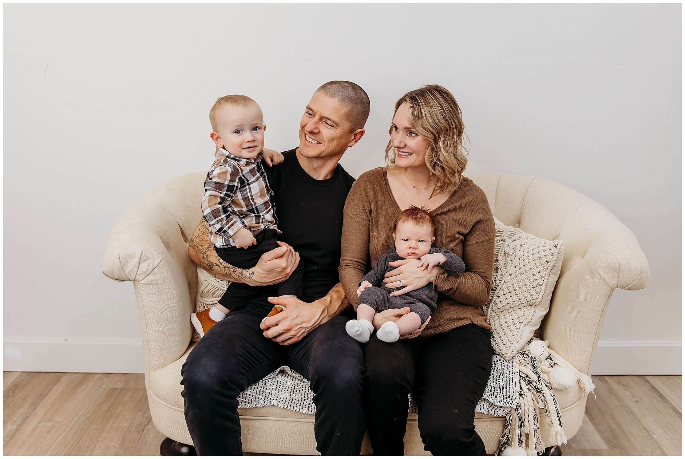 Family of four posing together in a downtown Everett photography studio during a lifestyle family session with baby Samson, older brother, and parents seated on a wicker chair.