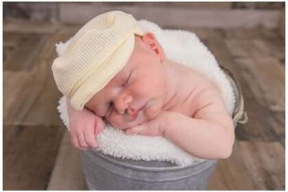 Sleeping newborn baby wearing a soft knit cap, curled up in a cozy blanket inside a small metal bucket, captured during a posed studio session.